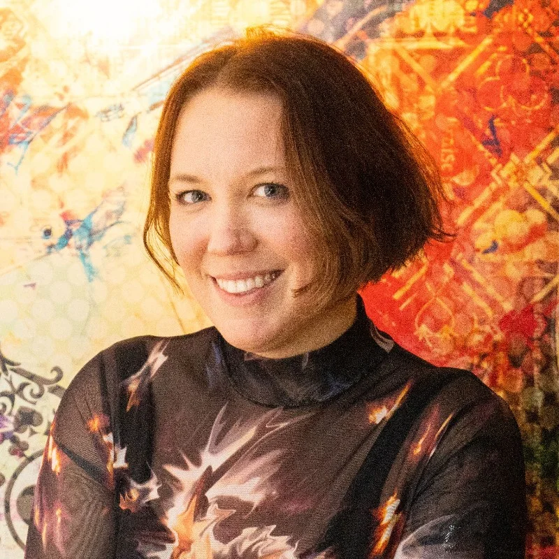 Liv's headshot. She has short reddish brown hair and is in front of a brightly colored wall