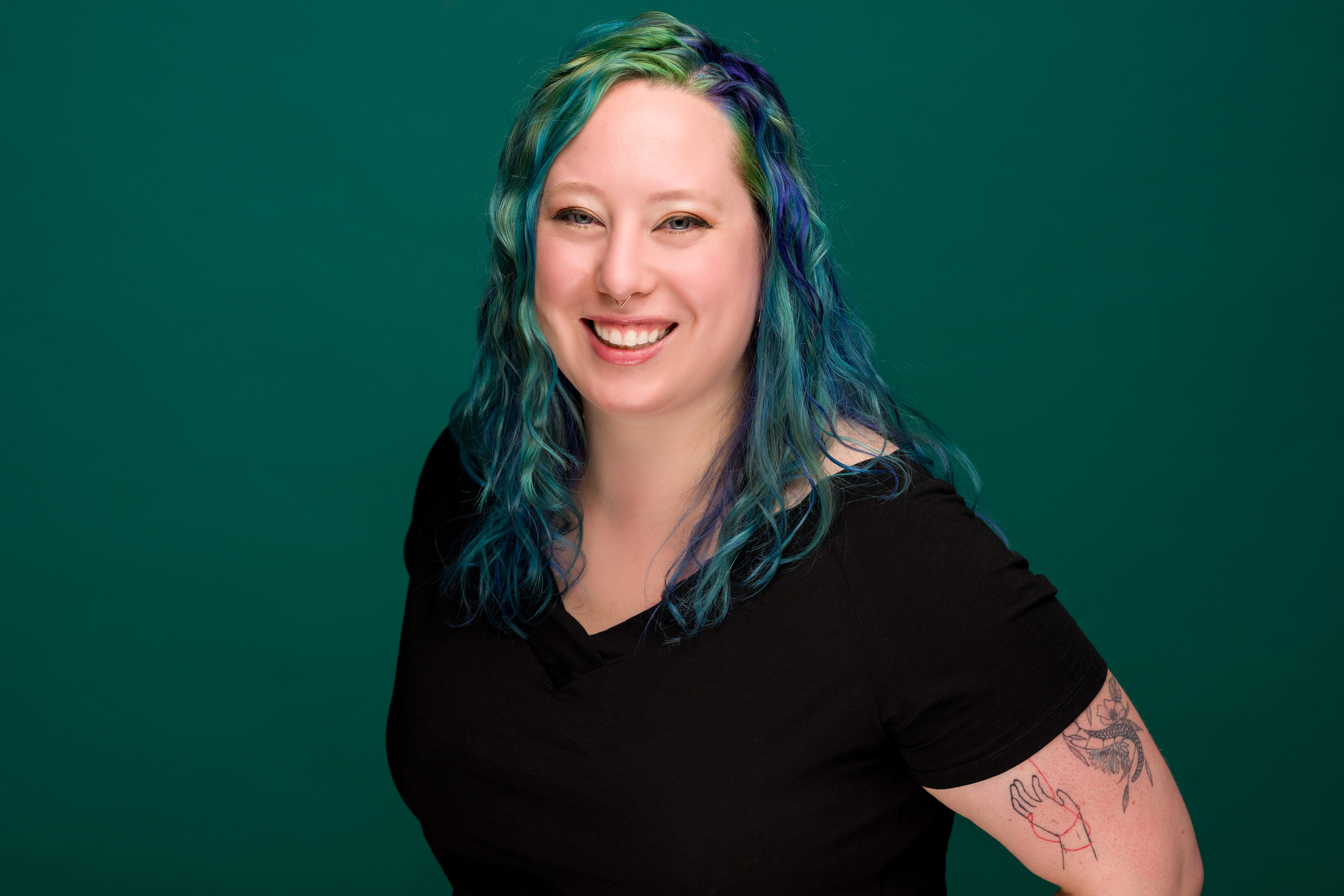 image of kat, a woman with blue and green hair, wearing a black shirt. she is angled toward the camera and smiling.