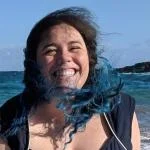 Tiffany, a white woman with curly blue and brown hair laughs in front of a blue ocean, and blue sky