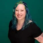 image of kat, a woman with blue and green hair, wearing a black shirt. she is angled toward the camera and smiling.